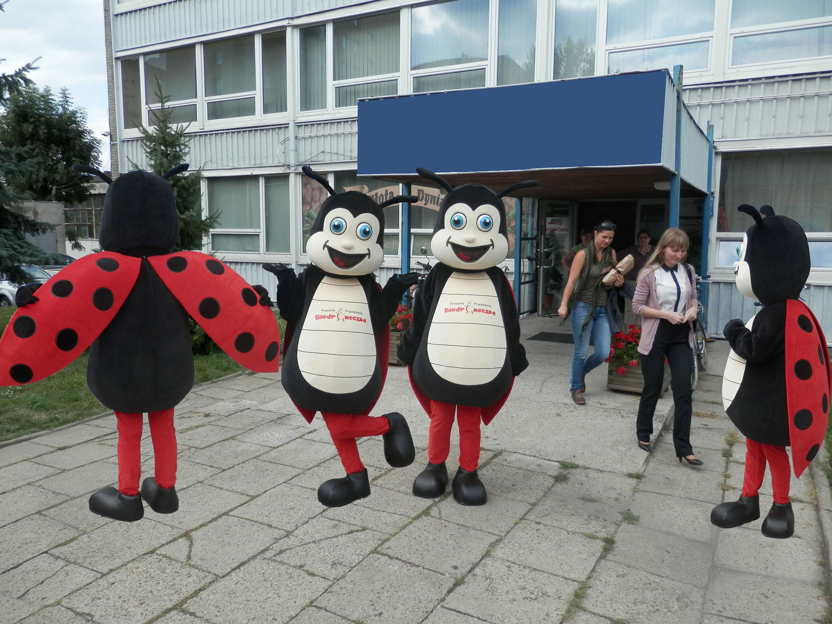 Na obrazku widnieje kostium biedronka, obok którego stoją strój biedronka, przebranie biedronka oraz maskotka biedronka. Fotografia wykonana przed siedzibą firmy Elabika. Obok maskotek widać również przechodzące osoby.